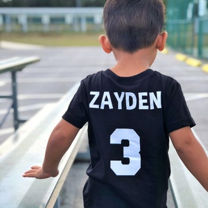 May include: A black t-shirt with the name "ZAYDEN" and the number "3" in white block letters on the back. The shirt is worn by a child standing near a bench.