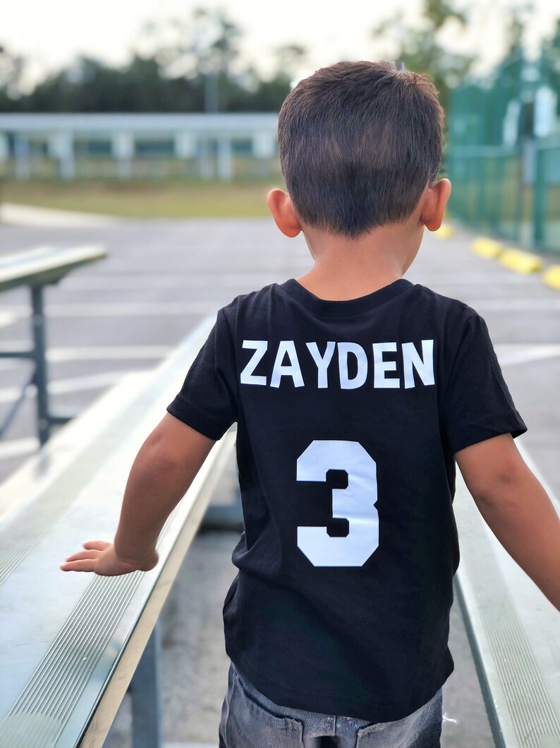 May include: A black t-shirt with the name "ZAYDEN" and the number "3" in white block letters on the back. The shirt is worn by a child standing near a bench.