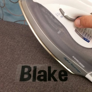May include: A white iron with a digital display is being used to press a clear vinyl sticker with the name "Blake" on a gray fabric.