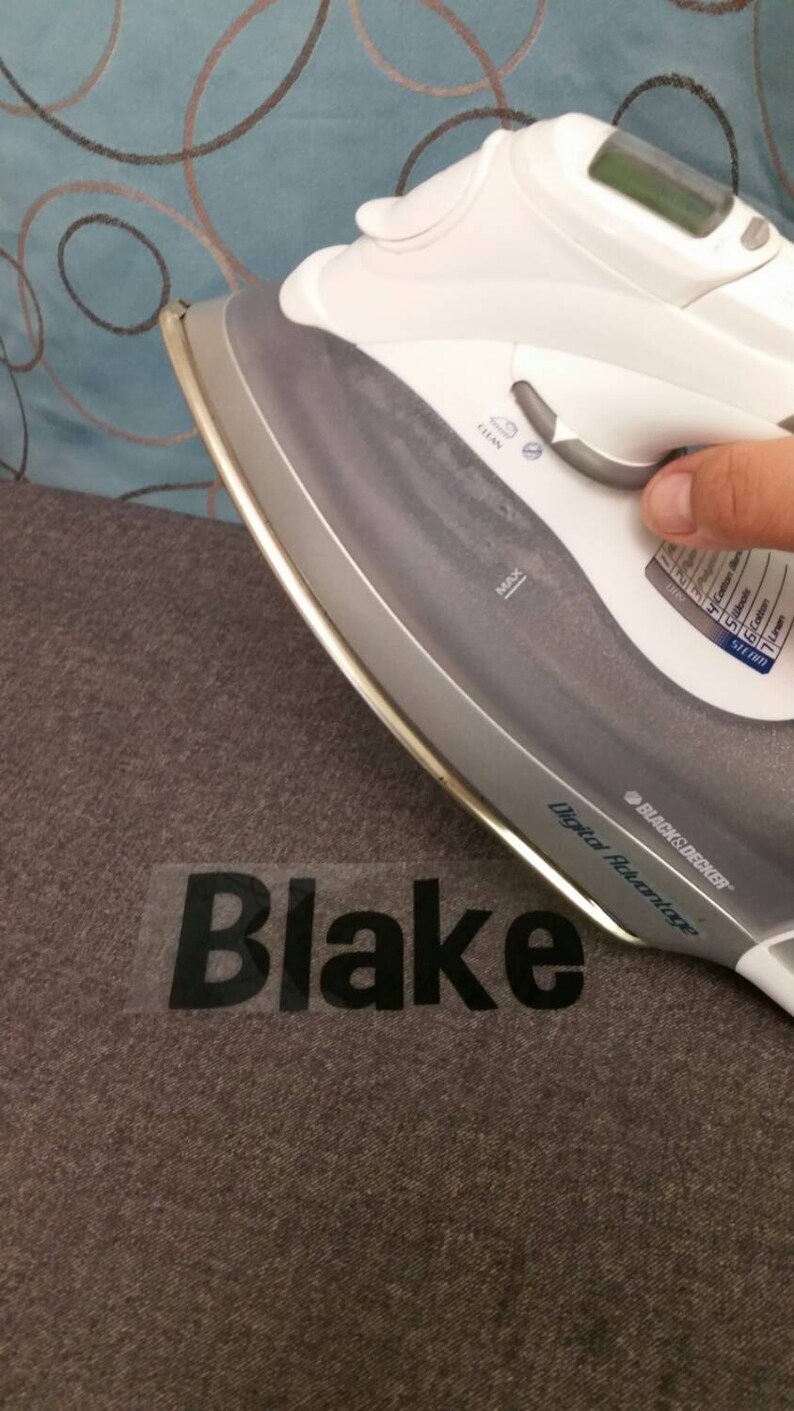 May include: A white iron with a digital display is being used to press a clear vinyl sticker with the name "Blake" on a gray fabric.