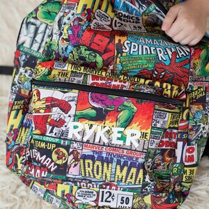 May include: A black backpack with a colorful comic book print featuring Marvel characters like Iron Man, Spider-Man, and the Hulk. The backpack has a white name tag that reads "RYKER".