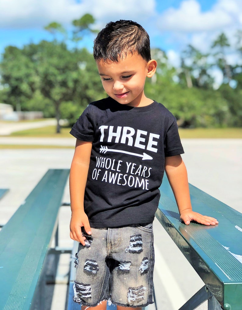 May include: A young child wearing a black t-shirt with the text "THREE WHOLE YEARS OF AWESOME" in white. The child is also wearing distressed denim shorts. The background features a park setting with a blue sky and trees.