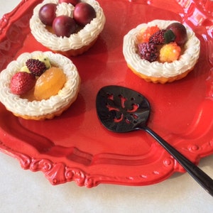 May include: Three mini fruit tarts with whipped cream and various fruits on a red plate with a black decorative serving spoon.