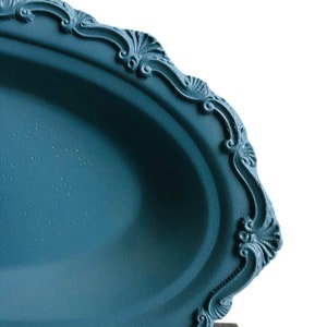 Può includere: Un vassoio ovale blu da portata con un bordo decorativo e ornato. Il vassoio è in metallo e ha una finitura liscia e opaca.