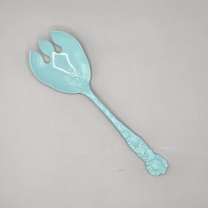 May include: A light blue plastic salad serving spoon with a floral design. The spoon has two prongs at the end and a rose-shaped handle.