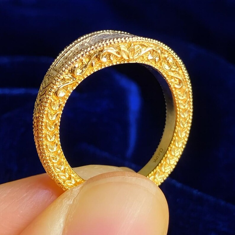 Puede incluir: Primer plano de un anillo de oro con intrincados patrones florales y de hojas. El anillo tiene una superficie texturizada y una banda con grabados detallados. El anillo se sostiene sobre un fondo azul oscuro.