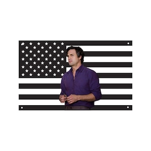 May include: A person wearing a purple button-down shirt stands in front of a black and white American flag. The flag has 50 white stars in the upper left corner and black and white stripes.
