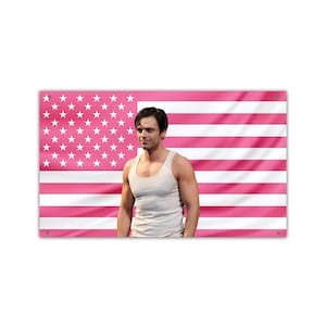 Puede incluir: Una bandera estadounidense rosa con estrellas y rayas blancas. Una persona con una camiseta sin mangas blanca está de pie frente a la bandera. La bandera es rectangular y tiene un fondo rosa con estrellas blancas en la esquina superior izquierda.