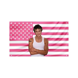 May include: A pink American flag with white stars and stripes. A person wearing a white tank top stands in front of the flag with arms crossed. The person has a tattoo on their left arm and is wearing a necklace and bracelet.