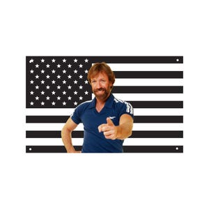 May include: A black and white American flag with a smiling person in a blue shirt pointing at the viewer. The flag has white stars in the upper left corner and black and white stripes.