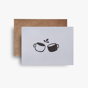 May include: A greeting card with a brown envelope. The card features a black and white illustration of two coffee cups clinking together, with coffee splashes above them. The card is on a white background.