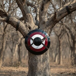 May include: A round shield with a black, red, and white design is strapped to a tree. The shield has a central black circle, a red band, and a white band. Brown leather straps secure the shield to the tree.