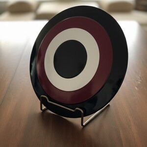 May include: A decorative plate with a concentric design in black, burgundy, white, and black, displayed on a metal stand. The plate has a glossy finish and a circular pattern. The stand is made of silver metal.
