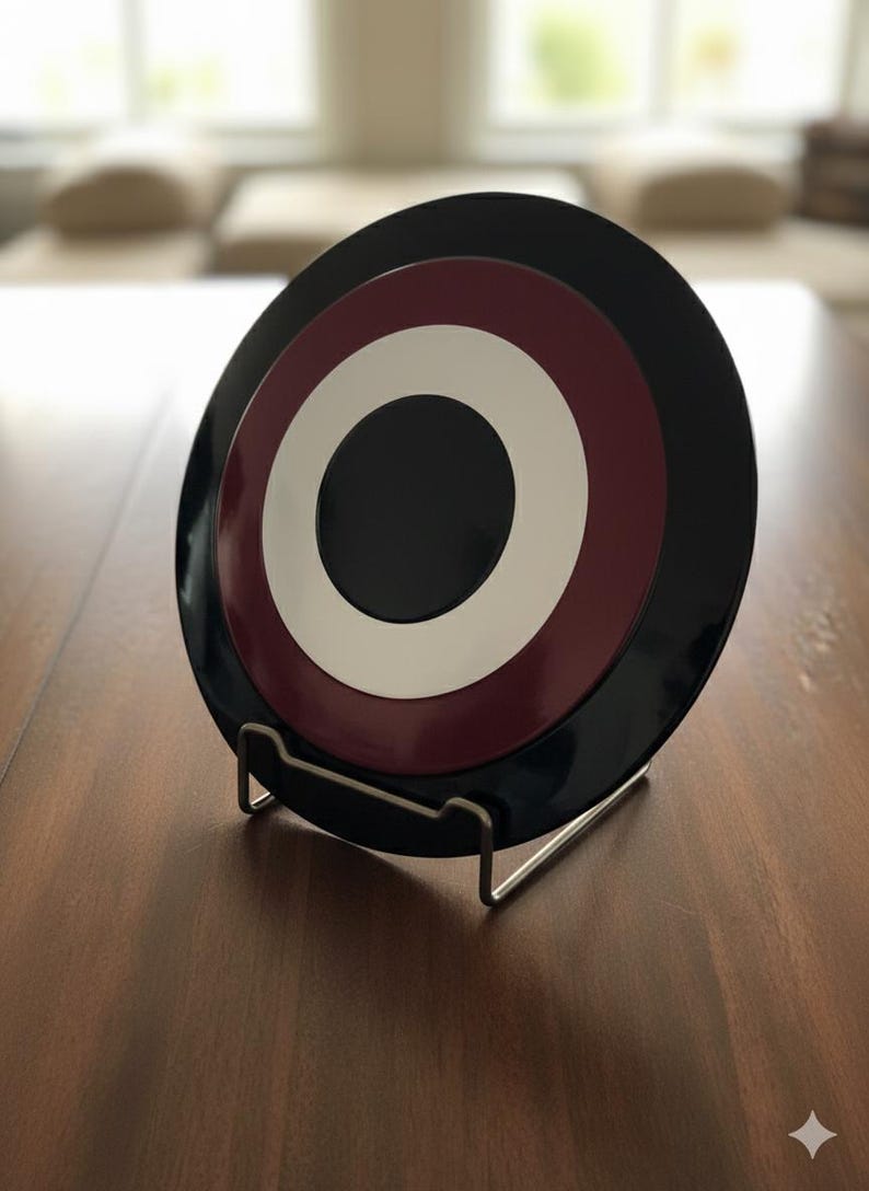 May include: A decorative plate with a concentric design in black, burgundy, white, and black, displayed on a metal stand. The plate has a glossy finish and a circular pattern. The stand is made of silver metal.