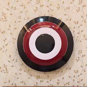 May include: A decorative wall art piece featuring a layered circular design. The piece has a black outer ring, a red middle ring, a white inner ring, and a black center. The artwork is mounted on a beige wall with a floral pattern.