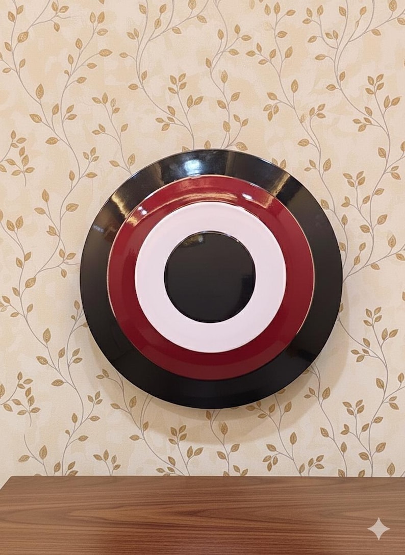 May include: A decorative wall art piece featuring a layered circular design. The piece has a black outer ring, a red middle ring, a white inner ring, and a black center. The artwork is mounted on a beige wall with a floral pattern.