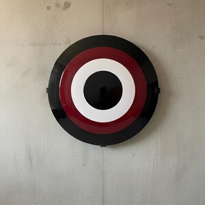 May include: A round, wall-mounted decorative piece with concentric circles in black, red, white, and black. The design resembles a target or a stylized emblem. The piece is mounted on a gray concrete wall.