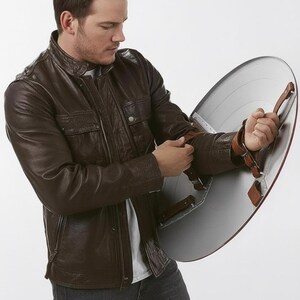 May include: A brown leather jacket with a zip-up front and a silver shield with brown leather straps. The shield has a red border and is held by the person in the image.