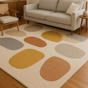 May include: A cream-colored area rug with large, irregularly shaped patterns in shades of yellow, orange, and gray. The rug is placed on a hardwood floor, and a gray sofa and wooden furniture are visible in the background.