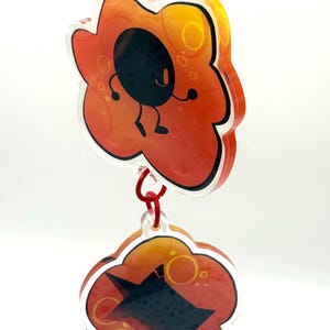 May include: Two-piece acrylic keychain with a red clasp. Each piece is a flower shape with a gradient orange and yellow background. One features a black character with a smiling face, the other a black shape.