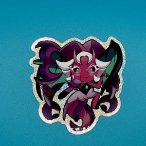 May include: A holographic sticker featuring a cartoon character with a red face, white hair, and purple accents. The character has large eyes and is surrounded by a glittery border. The sticker is on a blue background.