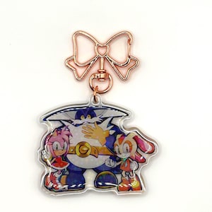 May include: A clear acrylic charm featuring the characters Big the Cat, Amy Rose, and Cream the Rabbit from the Sonic the Hedgehog series. The charm has a rose gold bow-shaped clasp. The characters are in vibrant colors.