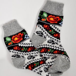 May include: Knitted socks with grey cuffs and heels. The socks have a white base with a black and red floral pattern. The cuff features a black band with red roses, green leaves, and blue accents. These socks are designed for warmth and comfort.
