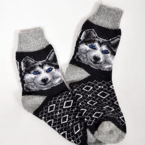 May include: A pair of black and grey socks with a detailed image of a husky dog with blue eyes. The socks have a diamond pattern on the foot and grey cuffs.