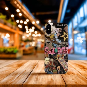 May include: A phone case featuring a collage of anime-style illustrations and the text "Soul Eater." The design includes characters, a skateboard, and a star on a black background. The case is on a wooden surface.