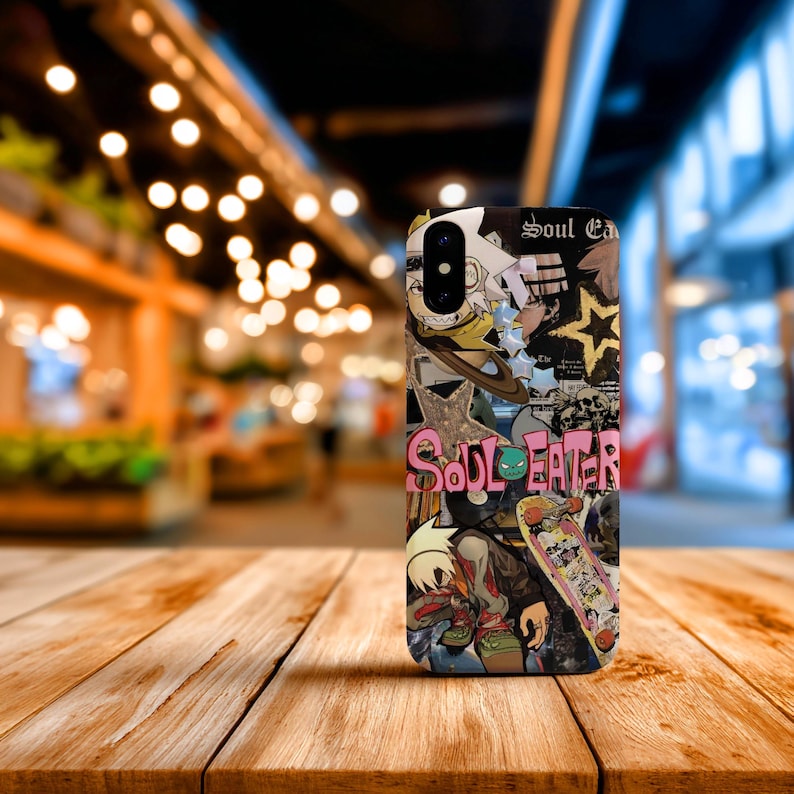 May include: A phone case featuring a collage of anime-style illustrations and the text "Soul Eater." The design includes characters, a skateboard, and a star on a black background. The case is on a wooden surface.