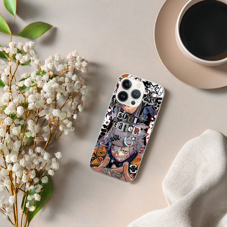 May include: A clear phone case featuring a colorful anime-style graphic with the words "SOUL EATER" in white. The design includes a character with gray hair and a sinister grin. The case is on a light beige surface with a cup of coffee and flowers.