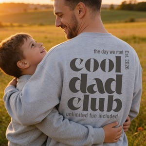 Könnte beinhalten: Ein hellgraues Sweatshirt mit Rundhalsausschnitt und dunkelgrauem Text auf der Rückseite, der the day we met Est. 2026 cool dad club unlimited fun included in einer fetten, abgerundeten Schrift lautet. Das Kleidungsstück hat lange Ärmel und einen gerippten Kragen.