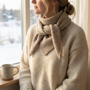 Può includere: Una sciarpa beige a maglia è drappeggiata attorno al collo di una persona che indossa un maglione abbinato. La sciarpa è annodata davanti, con le estremità che pendono. Una tazza di ceramica è appoggiata su un davanzale di legno. Lo sfondo mostra un paesaggio innevato.