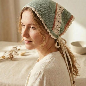 May include: A knitted head covering in shades of green, beige, and peach. The head covering has a scalloped edge and a patterned design on the side. Beige ties with wooden beads hang down the back. The head covering is likely handmade.
