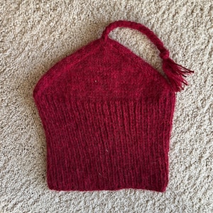 May include: A knitted red hat with a pointed top and a braided loop ending in a tassel. The hat is made of a textured yarn, possibly wool. The hat is a solid, deep red colour.