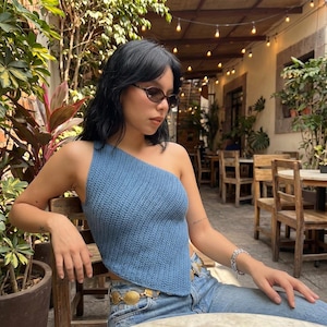 May include: A blue knitted one-shoulder top. The top is a solid light blue color and has a triangular shape. The model is wearing blue jeans and a gold belt. The background is a cafe setting with plants and wooden furniture.