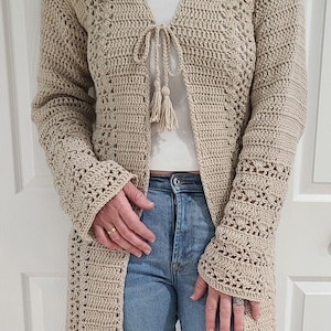 May include: A beige crochet cardigan featuring long sleeves with intricate openwork patterns, a V-neckline, and a front tie closure with two decorative tassels. The garment is crafted with a textured stitch pattern throughout the body and sleeves.
