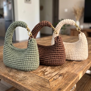 Può includere: Tre borse all'uncinetto, verde salvia, marrone e crema, sono appoggiate su un tavolo di legno. Ogni borsa ha un manico curvo fissato con elementi dorati. Le borse sono realizzate con un punto all'uncinetto stretto, che conferisce loro un aspetto strutturato.