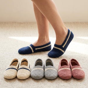 May include: Navy blue crochet slippers with a cream-colored stripe detail. Several pairs of crochet slippers in various colors, including cream, gray, and pink, are displayed on a neutral carpet. The slippers are designed for indoor wear.