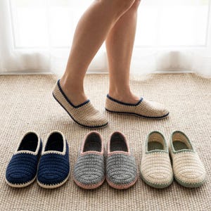 以下が含まれることがあります： さまざまな色の、かぎ針編みのスリッパがいくつか表示されています。スリッパには、ベージュ、ネイビーブルー、グレー、オフホワイトのオプションがあります。ベージュのスリッパはネイビーブルーのトリム、グレーのスリッパはピンクのトリムです。スリッパはニュートラルカラーのラグの上にあります。