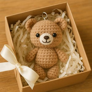 May include: A crocheted brown teddy bear keychain with black eyes and a white smile, presented in a brown gift box with a cream-colored ribbon. The box is filled with white paper strips.