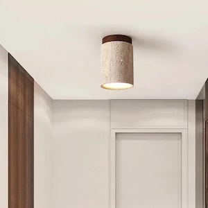 May include: A cylindrical ceiling light fixture with a textured, light-colored body and a dark brown top. The light is emitting a warm glow, illuminating the space below. The fixture is mounted on a white ceiling.