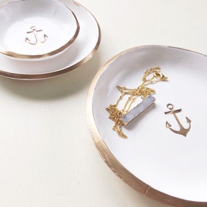 May include: White ceramic jewelry dishes with gold rims. One dish has a gold anchor charm, and another has a gold necklace with a blue-grey pendant. A smaller dish with an anchor is stacked on a larger dish.