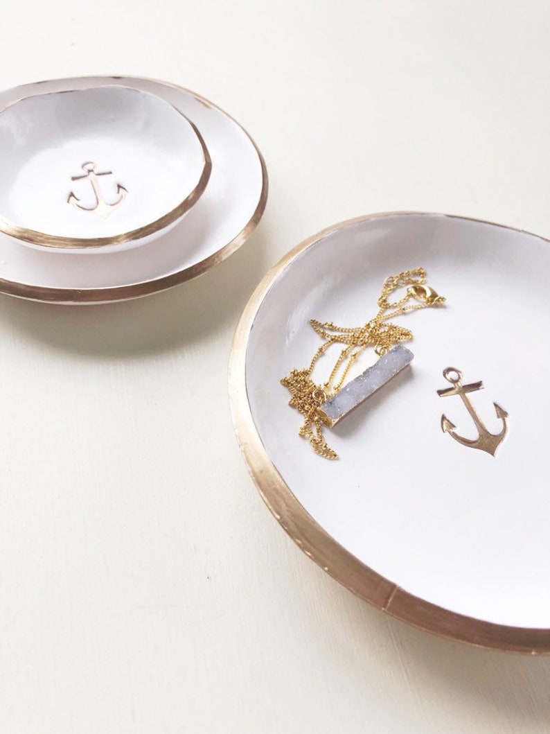 May include: White ceramic jewelry dishes with gold rims. One dish has a gold anchor charm, and another has a gold necklace with a blue-grey pendant. A smaller dish with an anchor is stacked on a larger dish.
