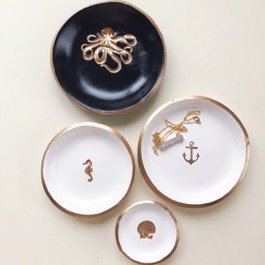 May include: Four decorative trinket dishes in varying sizes. The largest is black with a gold octopus, the others are white with gold rims and feature a seahorse, anchor, and seashell. A necklace rests on the anchor dish.