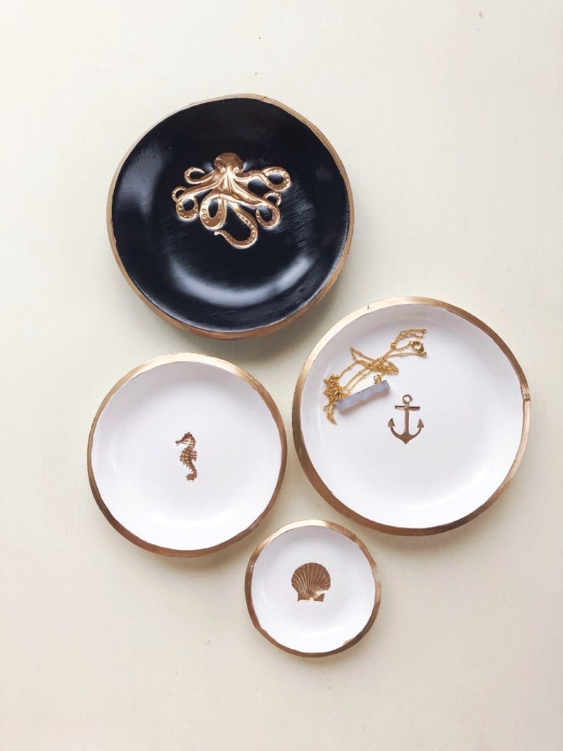 May include: Four decorative trinket dishes in varying sizes. The largest is black with a gold octopus, the others are white with gold rims and feature a seahorse, anchor, and seashell. A necklace rests on the anchor dish.