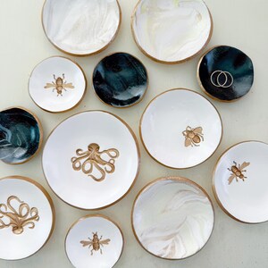 May include: A collection of decorative ceramic dishes in various sizes and designs. Some dishes feature gold rims and marbled patterns in white and teal. Others display gold illustrations of bees and octopuses. A few dishes are solid black.