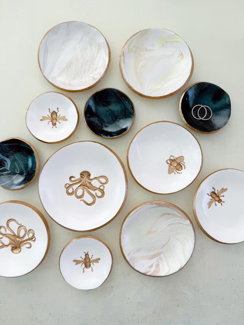 May include: A collection of decorative ceramic dishes in various sizes and designs. Some dishes feature gold rims and marbled patterns in white and teal. Others display gold illustrations of bees and octopuses. A few dishes are solid black.