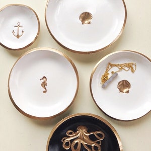 May include: A collection of decorative trinket dishes with gold rims. The dishes are white and black, featuring gold accents of an anchor, seahorse, seashell, and octopus. One dish holds a gold necklace. Ideal for nautical-themed decor.