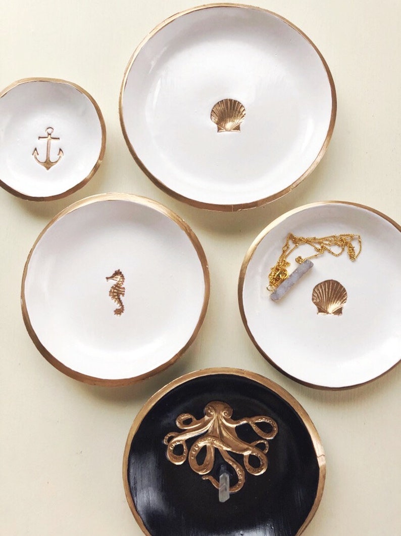 May include: A collection of decorative trinket dishes with gold rims. The dishes are white and black, featuring gold accents of an anchor, seahorse, seashell, and octopus. One dish holds a gold necklace. Ideal for nautical-themed decor.